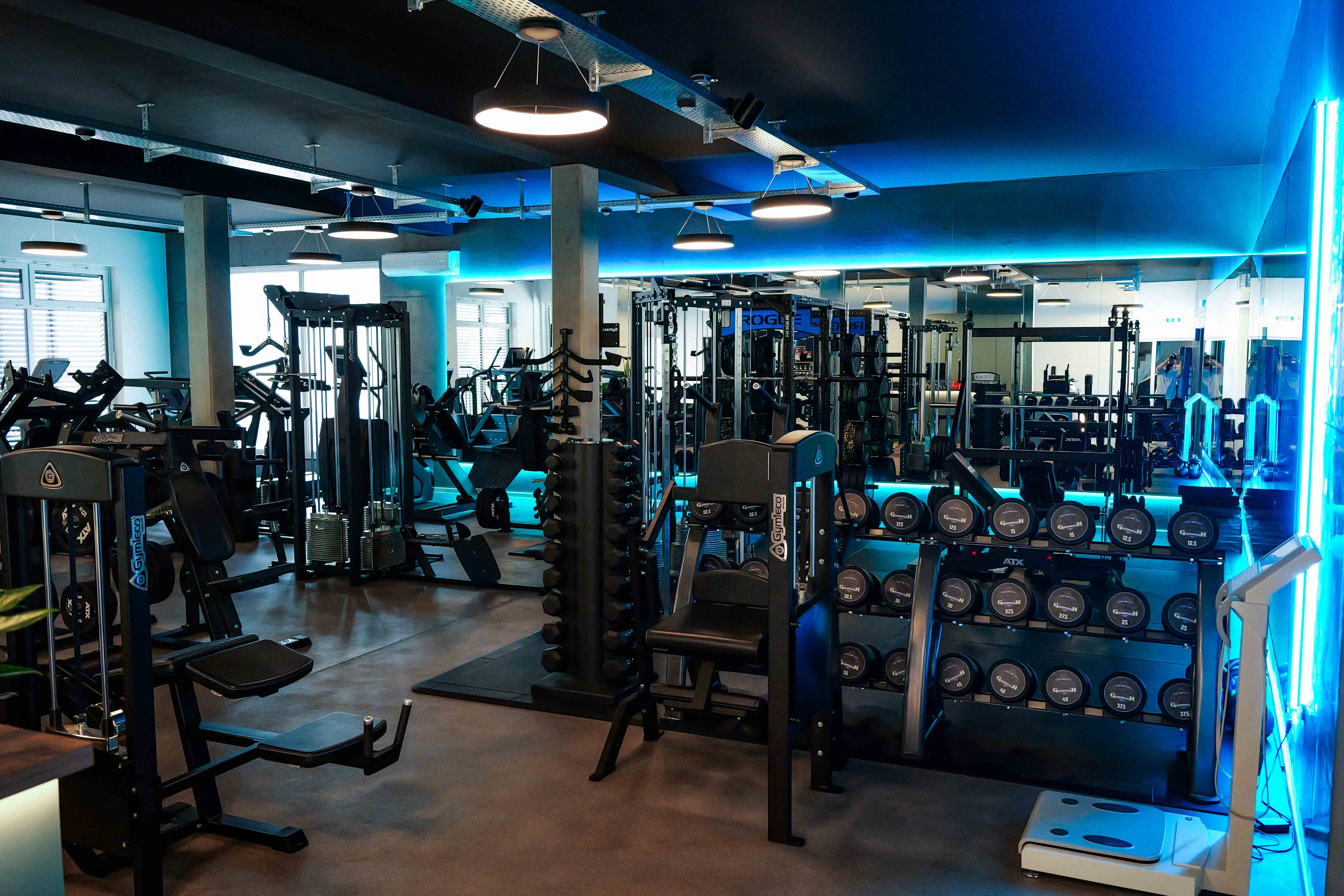 Innenansicht eines modernen Fitnessstudios mit Gewichten und Trainingsgeräten unter blauer Beleuchtung.