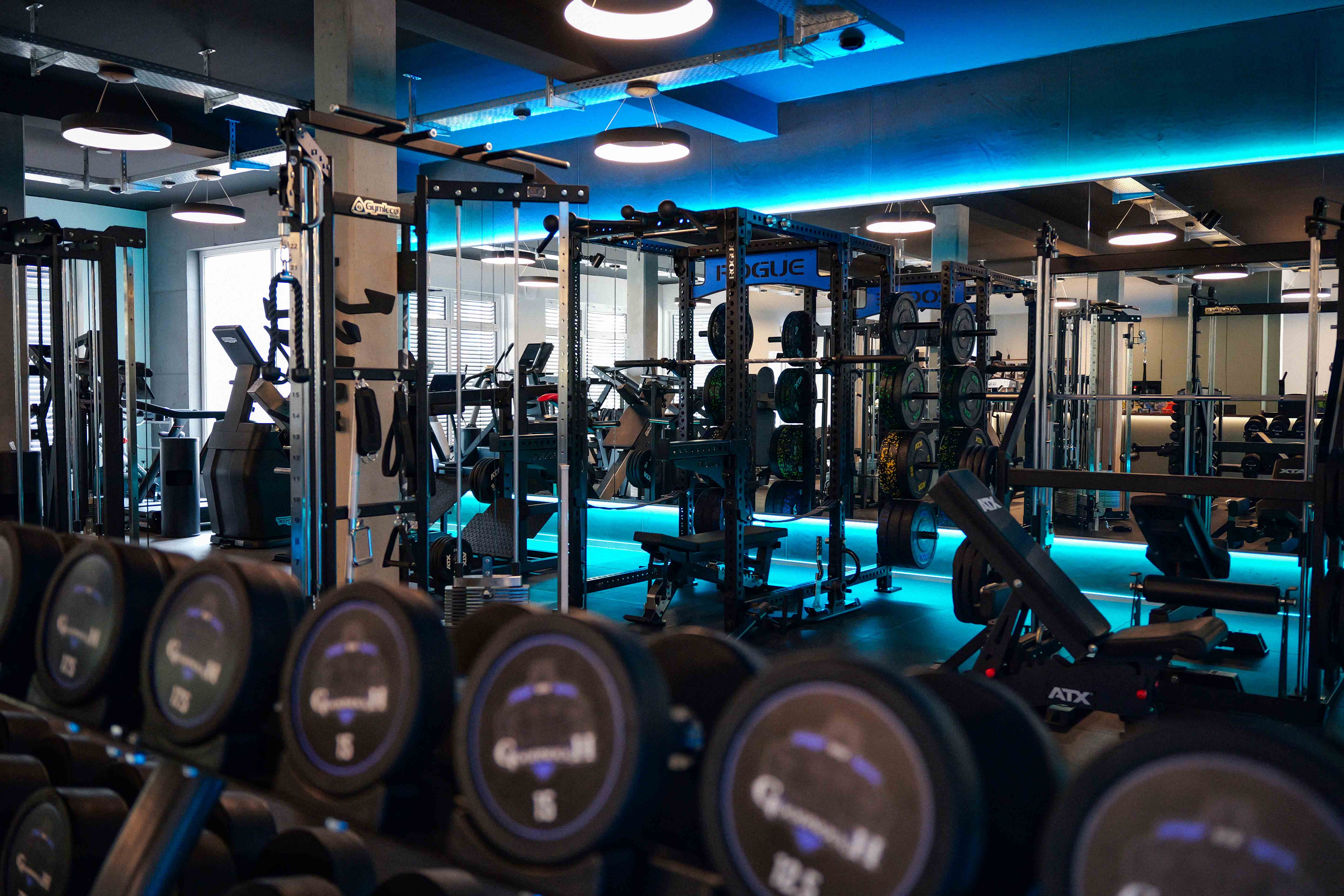 Moderne Fitnessstudio mit Kurzhanteln, Kraftstationen und Gewichten vor beleuchtetem Spiegel.
