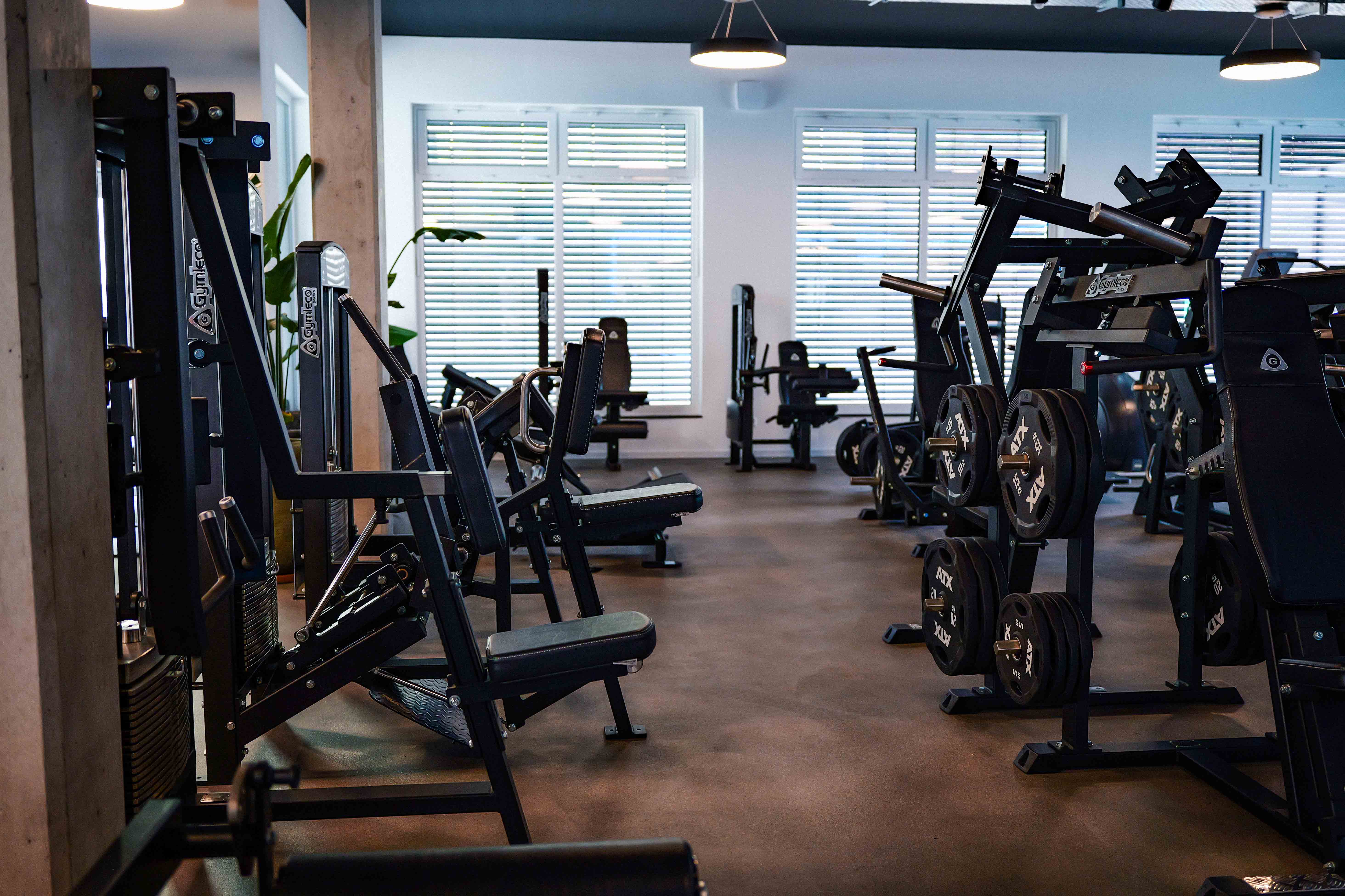 Moderner Fitnessraum mit diversen schwarzen Krafttrainingsgeräten und Gewichten an Fenstern mit Jalousien.