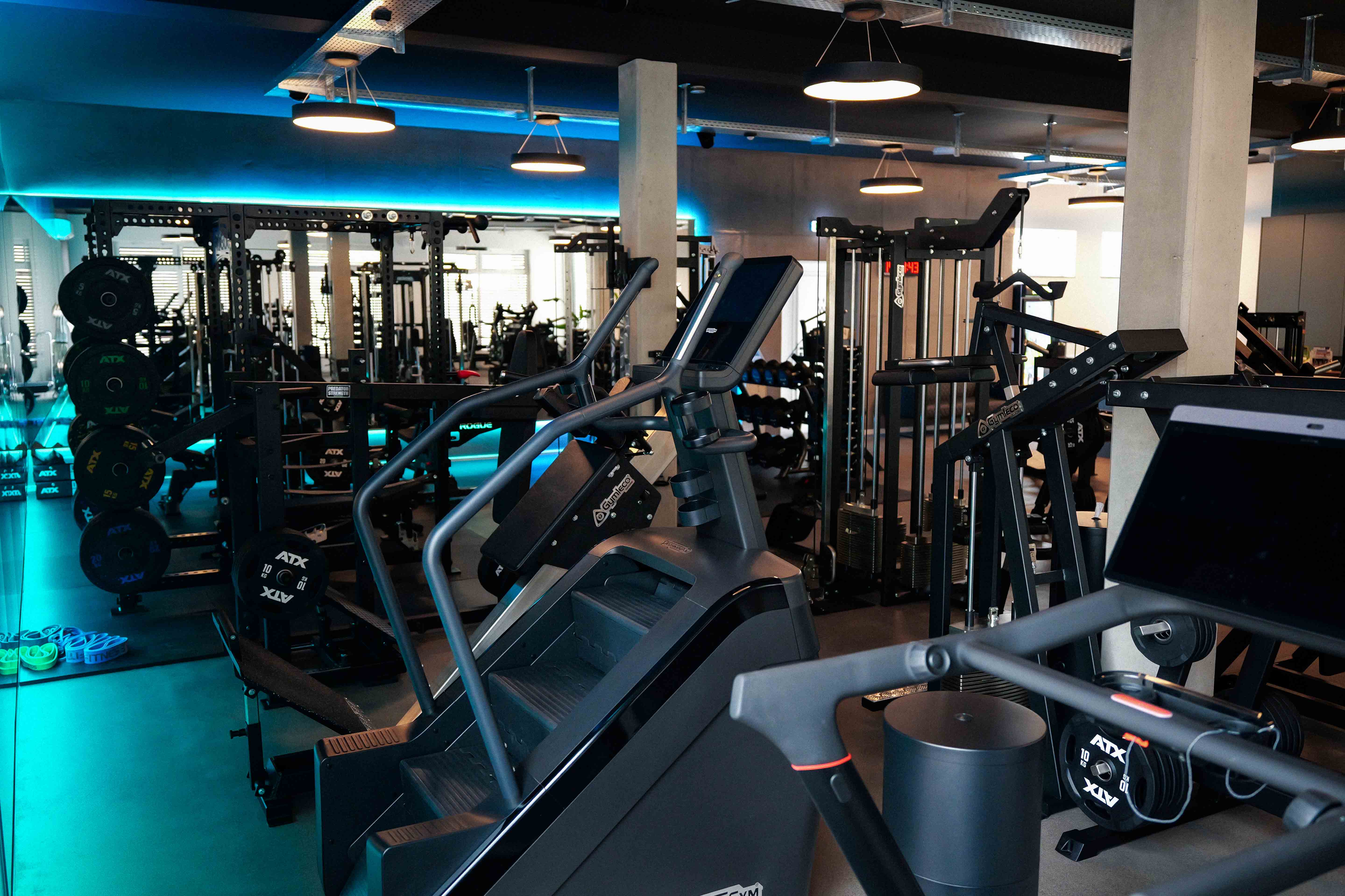 Moderne Fitnessstudio mit digitalen Trainingsgeräten, Hanteln und Gewichten in blau beleuchtetem Raum.