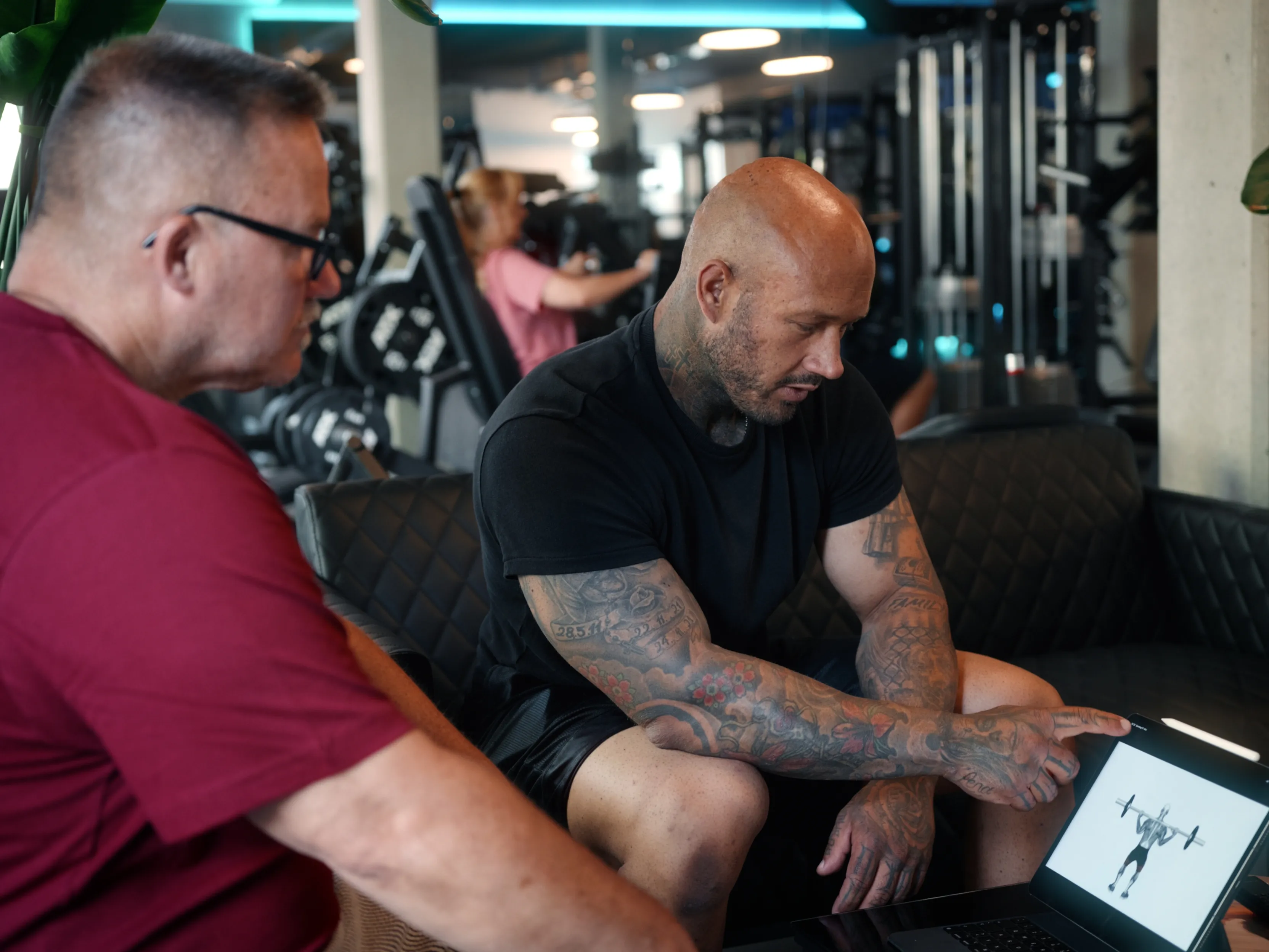 Zwei Männer im Fitnessstudio schauen auf Laptop mit Bild eines Mannes, der eine Langhantel hebt.
