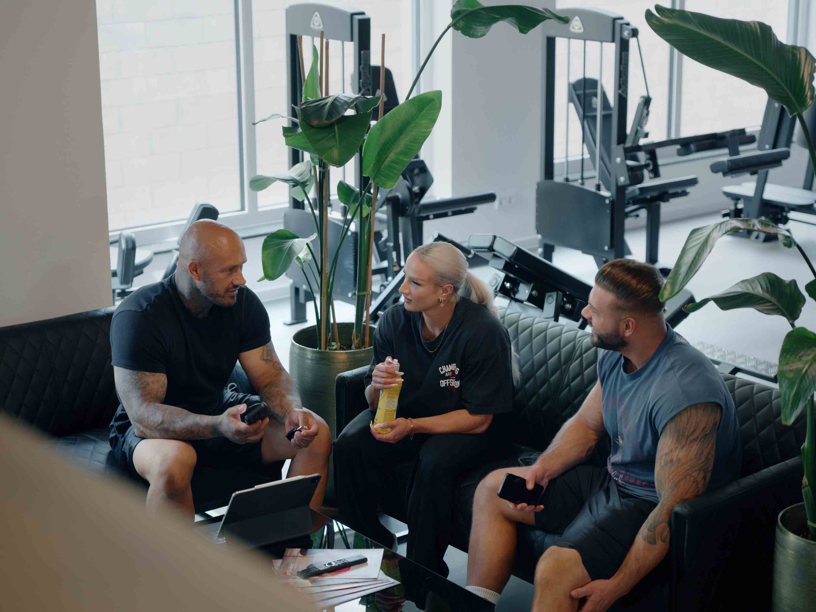 Drei Personen im Fitnessstudio sprechen, zwei Männer und eine Frau mit Getränk auf Ledersofas.