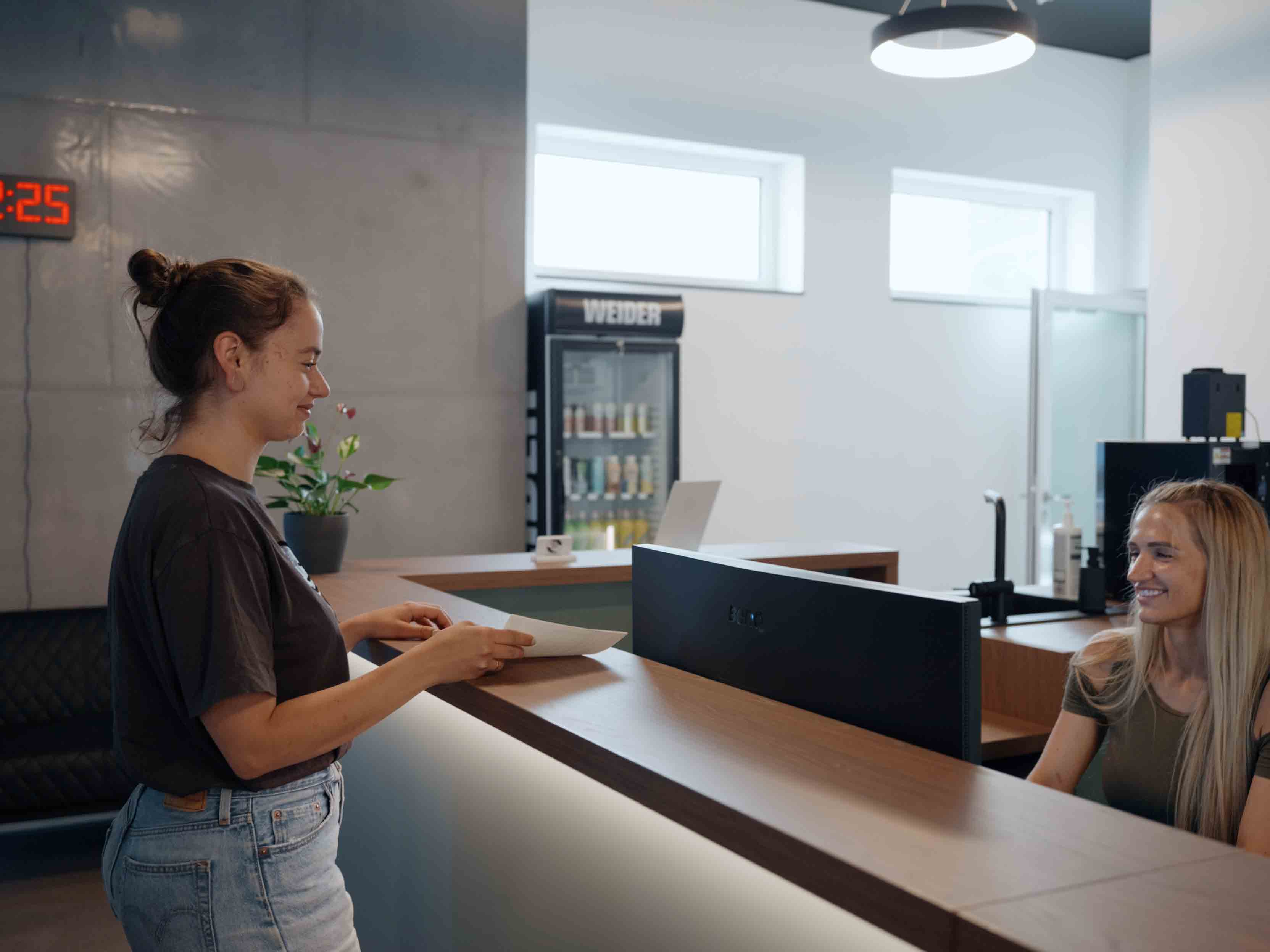 Frau an Empfangstresen spricht mit einer lächelnden Empfangsdame in modernem Büro.