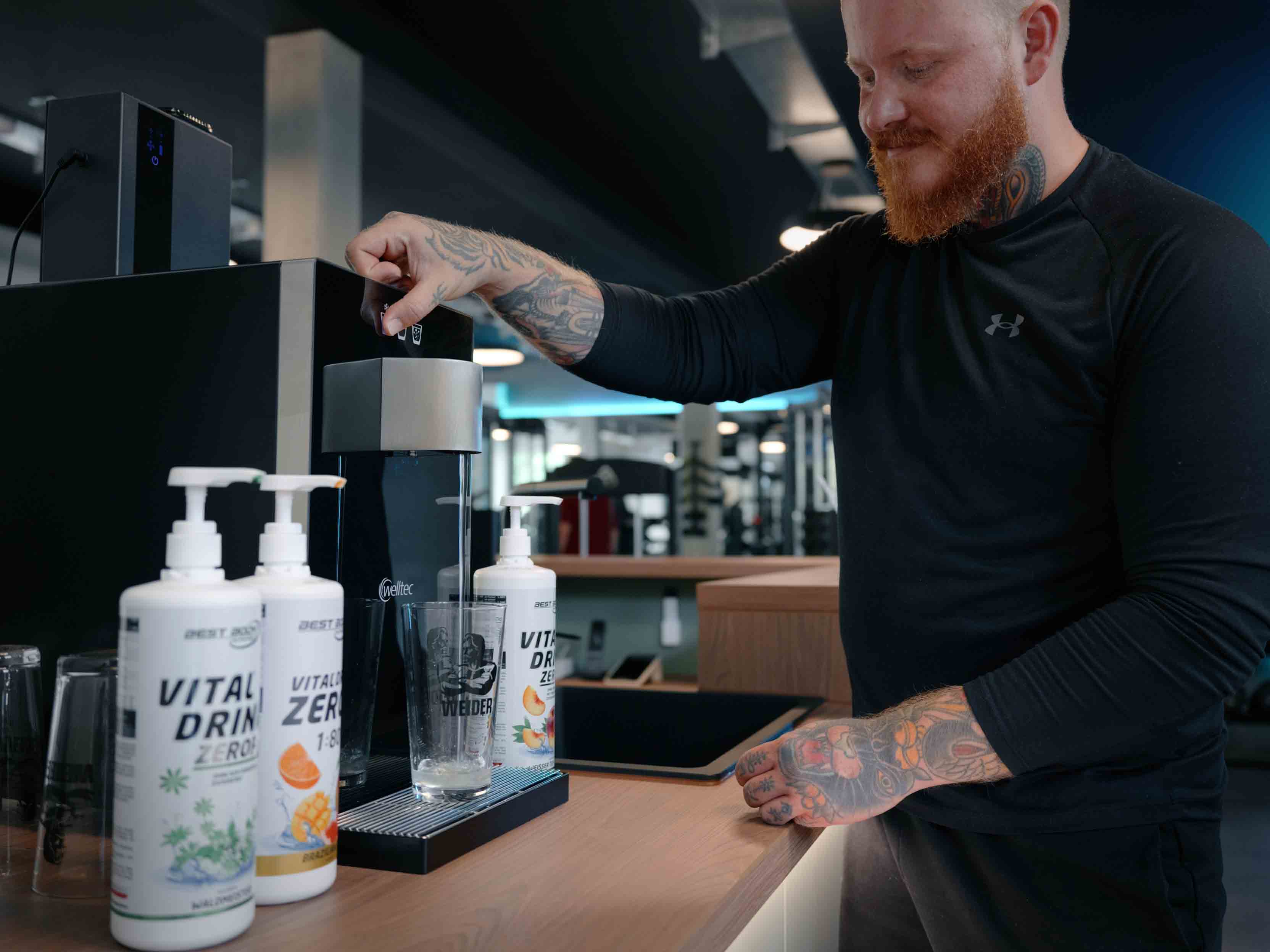 Mann mit Tattoos füllt in einem Fitnessstudio ein Glas an einem Wasserspender.