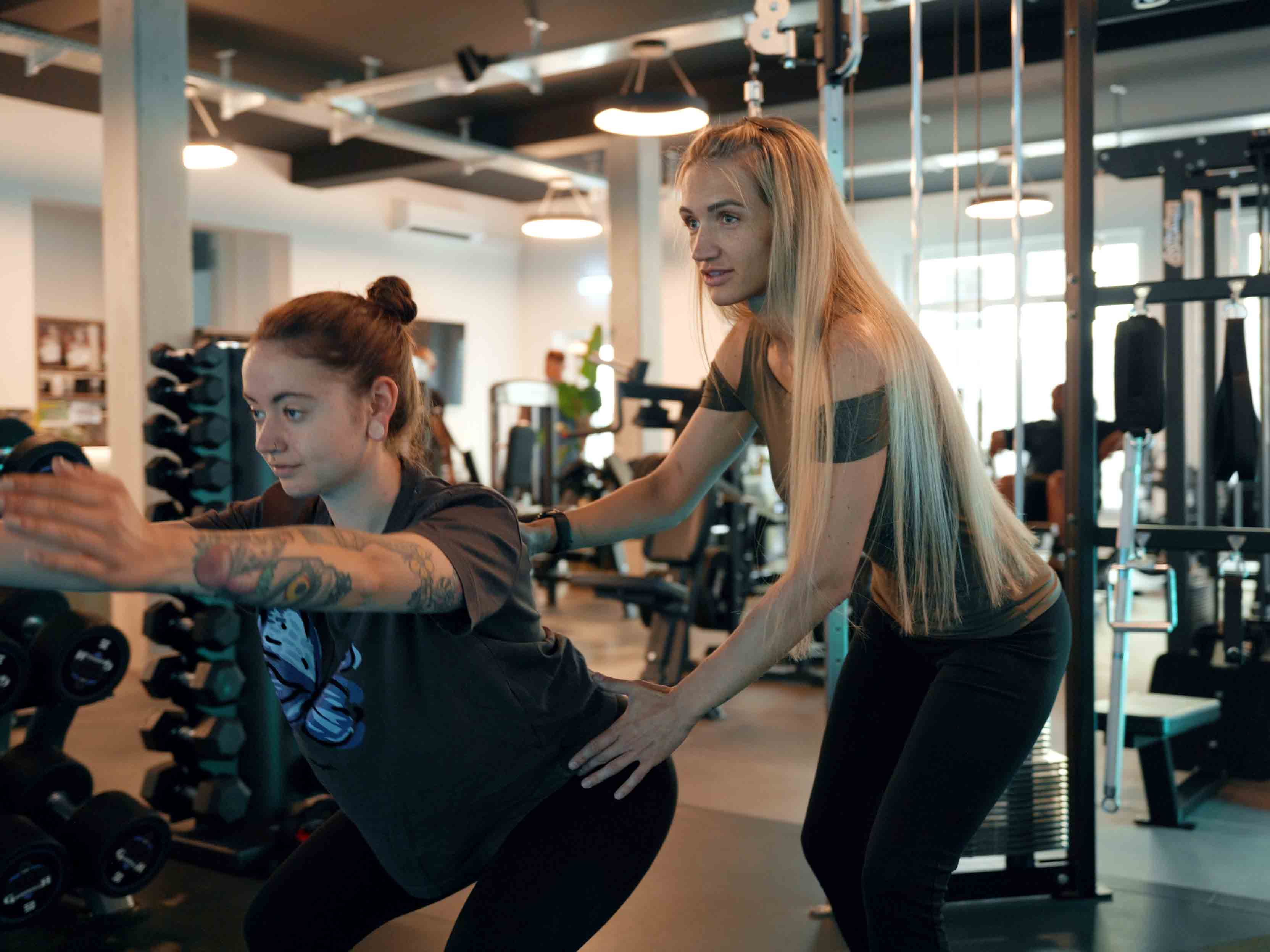 Trainerin hilft Frau mit Tattoo, beim Fitnesstraining im Fitnessstudio eine Übung korrekt auszuführen.